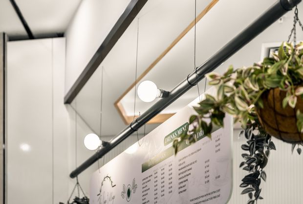 A close-up view of a contemporary café or commercial interior, featuring a white wall with a large illustrated menu board, black exposed pipe-style lighting rails, suspended round pendant bulbs, and hanging plants. A partial mirror or reflective surface and clean white cabinetry are visible in the background.