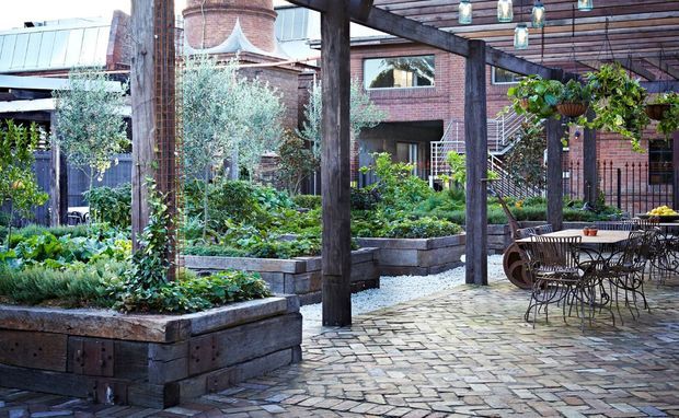 A landscaped courtyard garden with raised timber planting beds, mature greenery, a timber pergola, hanging planters, and a paved outdoor dining area. Exposed brick buildings and industrial-style structural elements surround the space.