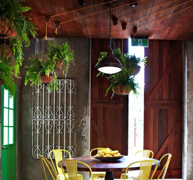 A rustic, atmospheric dining area with a dark timber-lined ceiling and large timber barn-style doors. A round dining table with yellow metal chairs sits beneath a hanging pendant light, while abundant suspended ferns and a decorative white wrought-iron grille add character. Green-framed glazing and an illuminated exit sign are visible in the background.