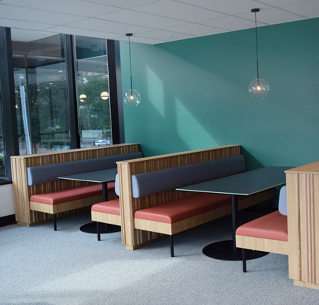 A modern commercial dining area with upholstered booth seating, timber slat partitions and bases, dark tabletops, pendant globe lights, teal walls, and large glazed windows. The space has a clean, contemporary café or restaurant fit-out with abundant natural light.