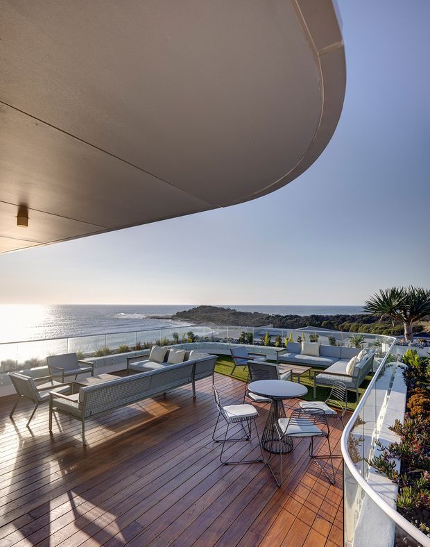 A spacious elevated outdoor terrace overlooking the ocean, furnished with multiple lounge seating areas and a small round dining table. The terrace has warm timber decking, glass balustrades, built-in curved white planter walls, and abundant coastal planting. A large curved overhead soffit or roof canopy frames the view, with bright sunlight and a calm seascape beyond.