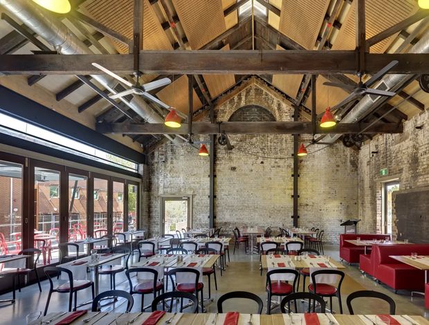 A spacious industrial-style dining hall or restaurant with exposed white-painted brick walls, heavy timber roof trusses, steel structural elements, exposed ductwork, acoustic wood ceiling panels, and polished concrete flooring. Numerous timber dining tables with black metal chairs and red upholstered seating are arranged throughout the room. Large glazed doors open toward an outdoor dining area, while red pendant lights and booth seating add colour and visual warmth.
