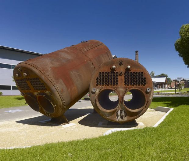 Outdoor photograph of a large weathered rust-colored industrial boiler or pressure-vessel sculpture displayed on a paved and gravel plinth in a landscaped public or commercial setting. The cylindrical metal form has riveted seams, circular openings, rows of perforations, and a tall chimney-like component. Modern low-rise buildings and manicured lawns surround the object under a clear blue sky.