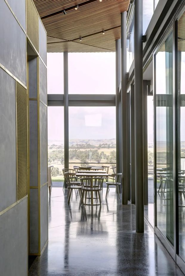A tall, modern glazed corridor or transitional interior opening onto an outdoor terrace, with floor-to-ceiling glass, slender grey structural columns, polished concrete flooring, and a slatted timber ceiling. Built-in wall panels with fine linear detailing line the left side, while café-style tables and chairs sit beyond the opening against a broad landscape view. The space has an airy, refined, institutional or commercial character with strong indoor–outdoor connection.