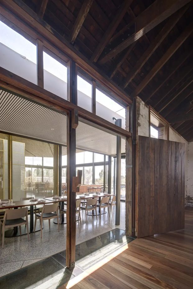 Interior view of a converted industrial-style dining or restaurant space, with exposed dark timber roof trusses and rafters, weathered timber posts and wall cladding, high clerestory glazing, and a glazed dining room beyond. The dining area contains neatly arranged tables and chairs, while polished concrete or terrazzo-like flooring and timber flooring define the foreground. Natural light enters through the extensive glazing and highlights the adaptive reuse character of the space.