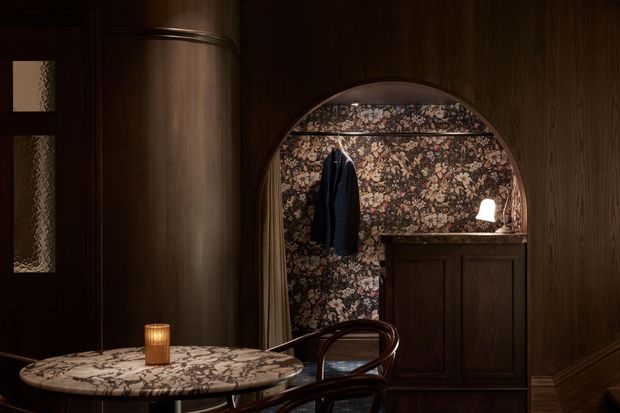A dark, intimate interior featuring rich dark timber wall panelling, an arched floral-patterned wallpaper alcove with a hanging coat, and a built-in timber cabinet. In the foreground is a round stone or marble-topped table with a candle and curved wooden chairs. The space has warm, low-level lighting and a refined, moody atmosphere.