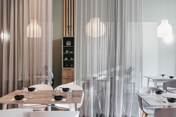 Bright contemporary dining area with multiple light timber dining tables, white molded chairs, sheer floor-to-ceiling curtains, pendant lighting, and a slim built-in shelving unit. A small adjoining dining or lounge area is visible through the curtains, creating a layered, airy restaurant-like interior.