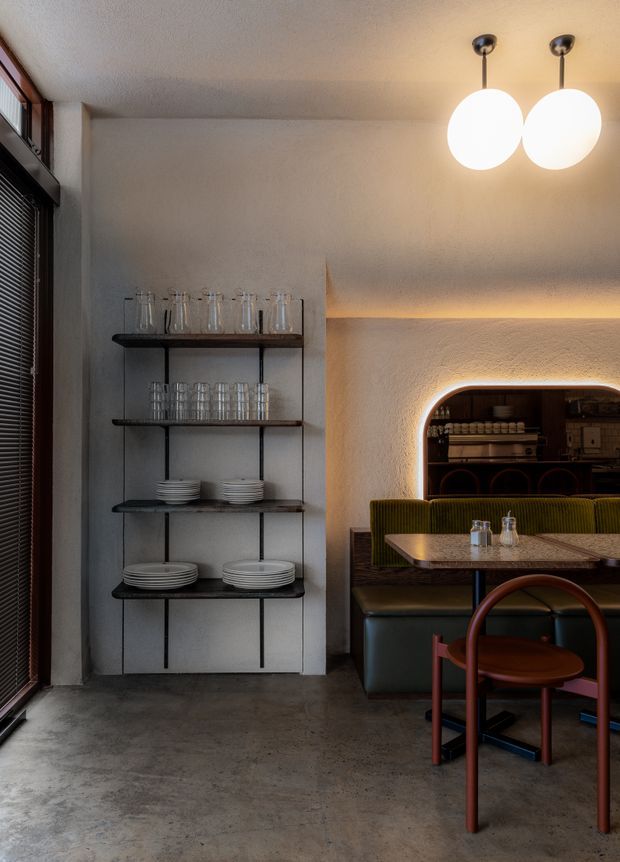 A warmly lit restaurant or café interior with an exposed concrete floor, textured plaster walls, open dark metal shelving displaying glassware and plates, and a built-in dining booth with olive-green upholstery. A rounded illuminated opening reveals additional bar or service joinery in the background. Globe pendant lights and a dark-framed window contribute to the refined contemporary atmosphere.