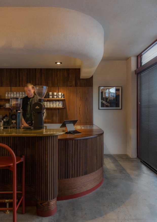 A compact, warmly lit coffee bar or café interior with a curved ribbed timber counter, dark timber wall panelling, integrated coffee equipment and shelving, a polished counter surface, and a concrete floor. A large curved plaster ceiling feature and dark-framed window with blinds add architectural character. The space has a refined, moody hospitality atmosphere with red seating accents.