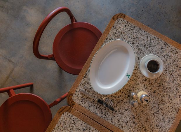 Overhead view of a richly textured terrazzo-topped dining table with rounded timber edging, set with a white oval plate, cutlery, a coffee cup, and metallic tableware. Deep red bentwood-style chairs surround the table, with a grey concrete floor beneath. The composition has a warm, moody, design-focused atmosphere.
