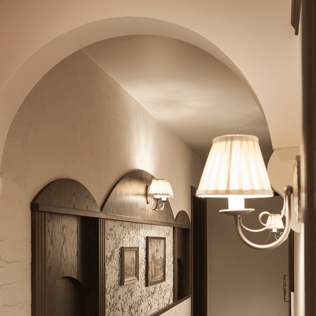 A warmly lit interior hallway or passage with a large curved plaster arch and barrel-vaulted ceiling. Dark timber arched wall panelling and doors frame a textured decorative wall covering with small framed artwork, while traditional wall sconces and a prominent cream fabric pendant-style light create a refined, atmospheric ambience.