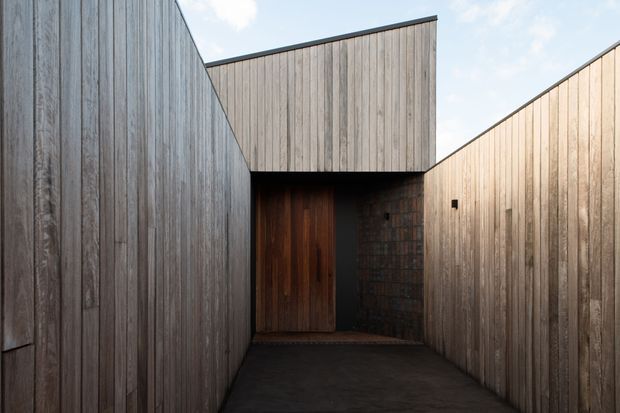 A narrow, enclosed entry passage leading to a dark timber front door. The passage is surrounded by tall vertical timber-clad walls and a projecting upper volume, with a small exposed brick section beside the entrance. The restrained palette and sharp geometric rooflines create a private, contemporary architectural facade.