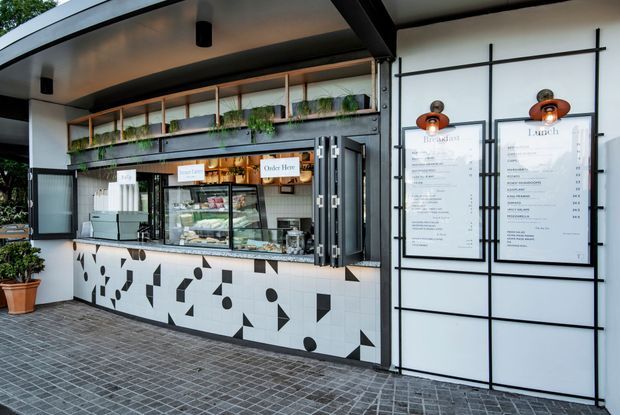 Exterior view of a contemporary food and beverage kiosk or café storefront. The open service counter has a bold black-and-white geometric tiled facade, displayed food and drinks, framed menu boards, black metal framing, timber shelving, and integrated planter greenery. The space fronts a paved outdoor area and uses a clean, graphic commercial fit-out.