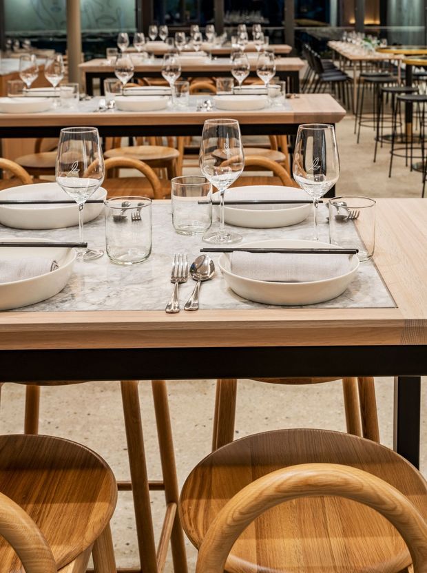 A warmly lit restaurant dining room with multiple formally set timber tables. The foreground table features a pale marble surface or inset, white ceramic bowls and plates, wine glasses, tumblers, and polished cutlery, surrounded by curved timber dining chairs. Additional tables and a bar-height communal setting recede into the background, creating a refined contemporary hospitality interior.