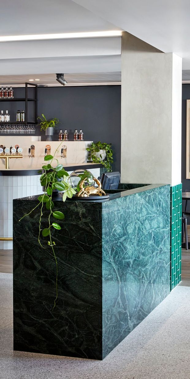 A dramatic contemporary home bar with a large sculptural island clad in dark veined stone on one side and blue-green textured stone on the other. The bar includes brass fixtures, a pale stone backsplash, dark shelving with glassware and bottles, and abundant trailing greenery. A dark feature wall, white ceiling elements, turquoise tile detailing, and terrazzo-style flooring complete the bold hospitality-inspired interior.