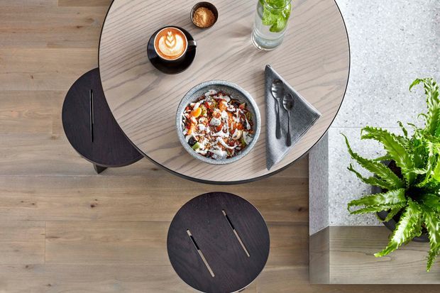 Overhead view of a compact dining area with a round light timber dining table, dark round stools, a bowl of food, coffee, water, and folded napkin with cutlery. The table sits beside a pale textured built-in surface and a lush fern, creating a warm, contemporary dining setting.