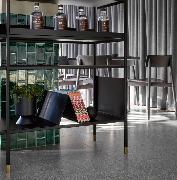A refined contemporary interior featuring a black metal open shelving unit with brass-toned feet, displaying bottles, books, and a potted plant. Behind it is a dining area with dark timber chairs and tables, grey floor-to-ceiling curtains, a green glass-block wall, and a polished grey floor. The space has a moody, sophisticated material palette with strong geometric lines.