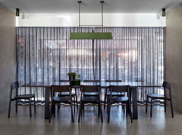 A contemporary dining room with a long dark timber dining table surrounded by matching chairs, positioned in front of full-height sheer pleated curtains. A suspended rectangular green pendant light hangs above the table, with potted plants as subtle table decor. The room has smooth neutral walls, a polished grey terrazzo floor, and a broad glazed opening behind the curtains.