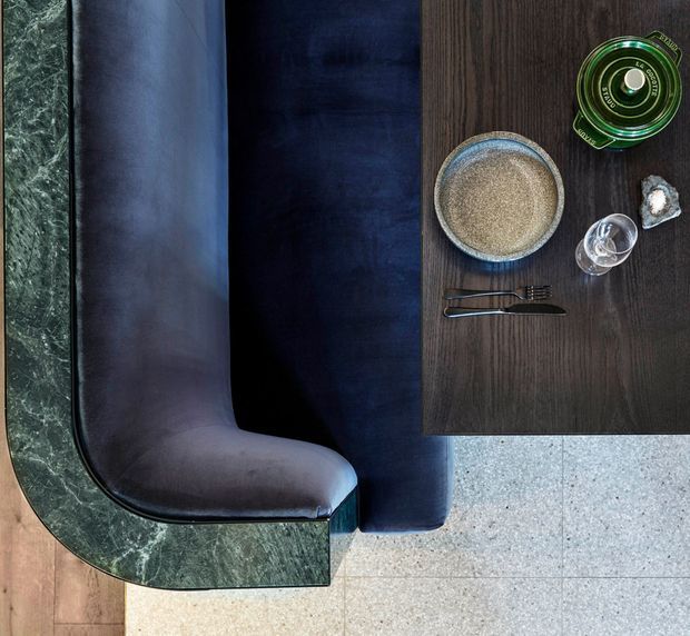 Overhead view of a refined dining or lounge setting featuring a dark upholstered curved chair or banquette, a green marble-topped rounded table edge, and a dark timber tabletop with a ceramic bowl, cutlery, glassware, and green glass vessel. The floor combines warm timber and pale stone or terrazzo-like paving.