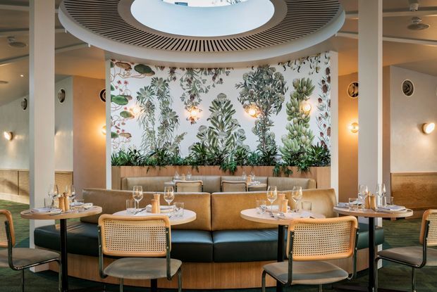 A refined restaurant dining area with a curved banquette, round dining tables, woven cane dining chairs, and warm ambient wall lighting. A botanical mural and integrated planter form a prominent feature wall, while a large circular ribbed ceiling detail with a central opening defines the space.