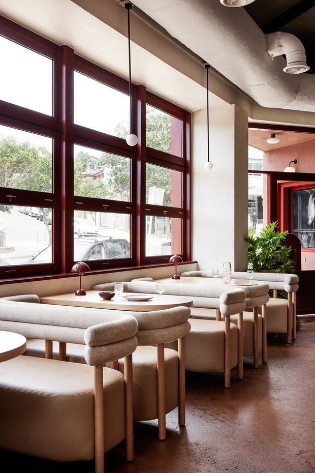 A warmly lit contemporary dining space, likely a restaurant or hospitality interior, with banquette seating and upholstered dining chairs arranged around pale wood tables. Large burgundy-red framed windows line one wall, while exposed ductwork, suspended globe pendants, and a polished earthy floor contribute to an industrial-influenced fit-out. A small indoor plant and service area are visible in the background.