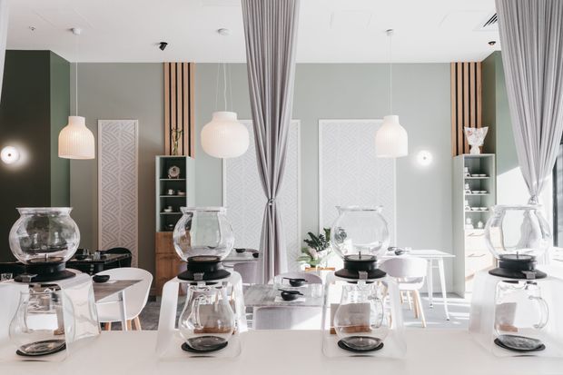 Bright contemporary dining area with multiple white tables set for coffee service, surrounded by white dining chairs. The room features pale grey walls, a dark green accent wall, vertical timber detailing, sculptural white pendant lights, patterned wall panels, open shelving, and full-height sheer curtains dividing the space.