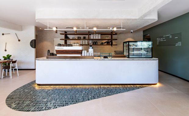 Contemporary café or hospitality counter with a large curved white service island, dark mosaic floor inlay, timber open shelving, display cases, pendant lighting, and a dark green feature wall with signage. A small seating area is visible to the left.