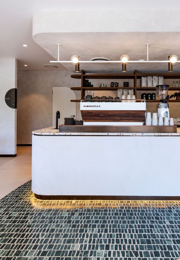 A stylish café or coffee bar interior featuring a curved white service counter, dark timber open shelving, espresso equipment, and warm pendant lighting. The counter has a richly veined stone or terrazzo-like benchtop and wood-clad front, while the floor is finished in a distinctive small-format dark blue-grey and cream mosaic tile pattern. The space has a warm, contemporary fit-out with textured plaster walls and rounded architectural forms.