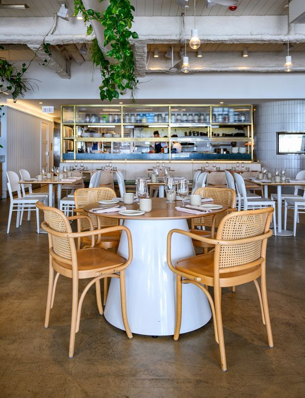 Bright contemporary restaurant interior with a central round dining table surrounded by woven cane chairs, polished table settings, and multiple rows of white dining tables. A glass-fronted service counter and open kitchen occupy the rear wall, framed in brass-toned metal and set against white tiled walls. The ceiling exposes insulated services and ductwork, while hanging greenery and pendant lights add warmth.