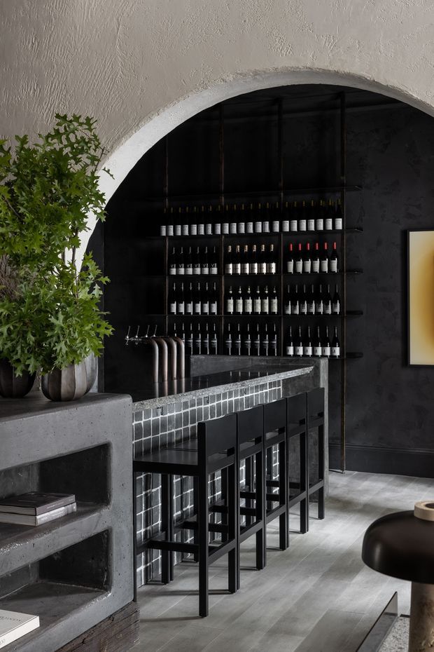 A dark, sophisticated home bar beneath a large textured arched opening. Black wall shelving displays rows of bottles and glassware behind a tiled bar counter with black bar stools. The space also includes a substantial grey concrete built-in shelving unit, dark wall finishes, grey flooring, and potted greenery, creating a moody, refined atmosphere.