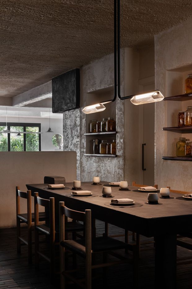 A moody contemporary dining area with a long dark dining table, black timber-framed chairs, neutral ceramic tableware and small candles. The space features textured plaster walls and ceiling, recessed display shelving with glass jars, exposed black conduit, sculptural linear wall lights, and dark timber flooring. A kitchen or service area is visible beyond a partial dividing wall, with large windows providing greenery-filtered daylight.
