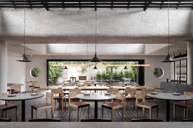 A contemporary restaurant dining room viewed from the rear, featuring multiple round and rectangular dining tables, simple wooden chairs, dark tiled flooring, and a long upholstered banquette along the right wall. Black industrial-style pendant lights hang from an exposed dark ceiling structure, while textured white walls and a broad glazed opening connect the dining area to greenery outside. Circular wall mirrors and restrained timber-and-grey finishes complete the refined hospitality interior.