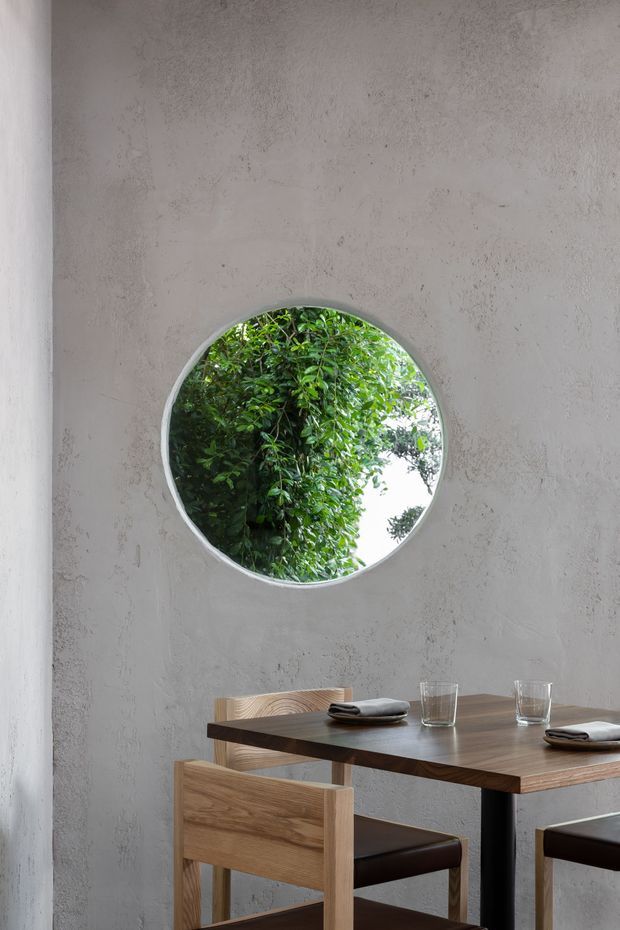 A minimalist dining area with a dark timber rectangular table and simple timber dining chairs, set against a textured pale grey plaster wall. A circular wall opening frames dense greenery outside, creating a strong natural focal point. The restrained palette and sparse furnishings give the space a calm, refined atmosphere.