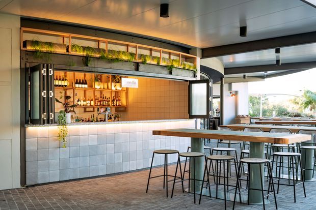 A contemporary open bar and dining area with a tiled service counter, illuminated bottle shelving, hanging greenery, timber-topped communal tables, and black metal stools. The space has a covered outdoor or semi-outdoor feel, with a clean linear ceiling, extensive openings, and bright natural light beyond.