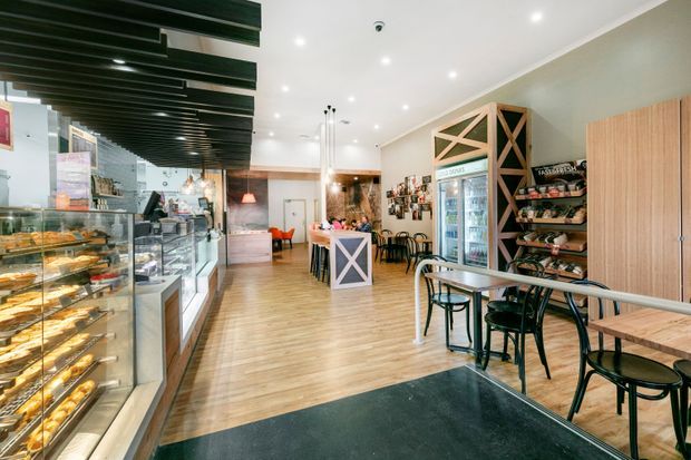 Bright, contemporary café or bakery interior with long illuminated pastry display cases, a service counter, timber-look flooring, black ceiling battens, pendant lighting, wall shelving, refrigerated drinks display, and café seating along the right side.