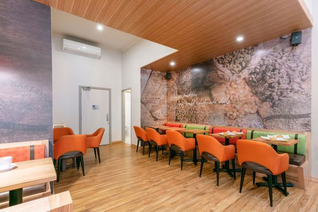 A contemporary dining area in a commercial or hospitality setting, featuring rows of orange upholstered chairs and tables, a large photographic feature wall, timber-look flooring, and a warm timber slatted ceiling. White walls, recessed lighting, wall-mounted speakers, and an air-conditioning unit complete the space.