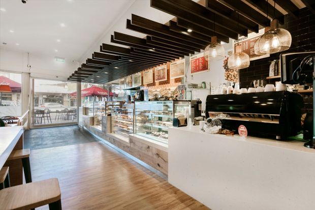 Bright contemporary café interior viewed along a long service counter, with glass pastry and food display cases, a large commercial coffee machine, tiled feature wall, pendant lights, framed menu boards, timber-look flooring, and exposed dark timber ceiling battens. Full-height glazing at the front opens views to an outdoor seating area.