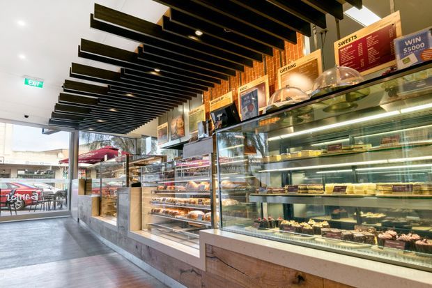 A bright contemporary dessert and café retail interior with long glass display counters filled with cakes, pastries, and sweets. The space features timber-clad counter fronts, extensive glazing, black linear ceiling battens, recessed lighting, menu boards, and outdoor seating visible through the storefront.