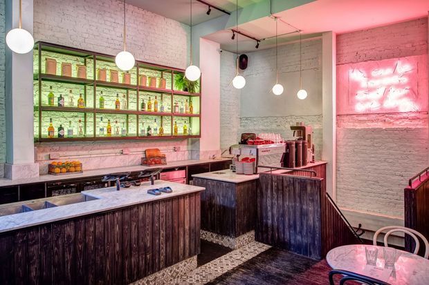 A stylised industrial bar or café interior with dark timber-clad counters, pale marble or stone benchtops, exposed white-painted brick walls, open back-bar shelving stocked with bottles, pendant globe lights, track lighting, and pink neon illumination. The space has a compact service area and a small seating zone in the foreground.