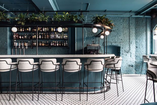 A moody, industrial-style bar interior with a long curved counter, upholstered high stools, exposed blue-painted brick walls, dark metal shelving stocked with bottles, pendant globe lighting, and suspended greenery. Patterned tile flooring and dark teal finishes create a refined hospitality setting.