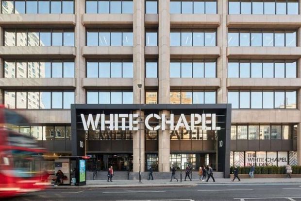 Street-level exterior view of a multi-storey commercial building with a regular grid of large glazed windows, stone or concrete vertical piers, and a prominent black entrance canopy marked “WHITE CHAPEL.” Pedestrians and a passing bus appear in the foreground.