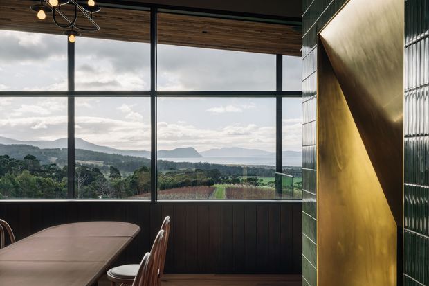 A refined dining area with a long table and chairs beside expansive black-framed floor-to-ceiling glazing. The room features dark vertical timber wall panelling, a warm timber ceiling, a sculptural black pendant light, and a dramatic metallic gold angular feature wall. The glazing overlooks rolling green hills, distant mountains, and agricultural land. The overall design combines warm natural materials with bold contemporary detailing.