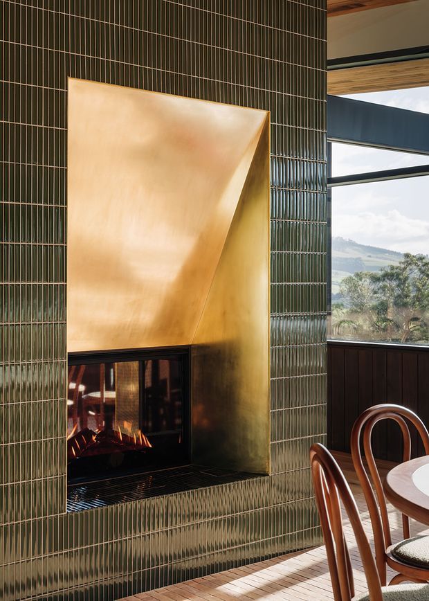 A warm, refined dining area centred on a built-in fireplace. The fireplace surround and adjacent wall are clad in narrow glossy dark green tiles with fine vertical and horizontal grout lines, while the firebox has a sculptural polished brass or copper-toned hood. A large black-framed window overlooks a hilly landscape, and curved timber dining chairs and part of a round dining table are visible in the foreground.