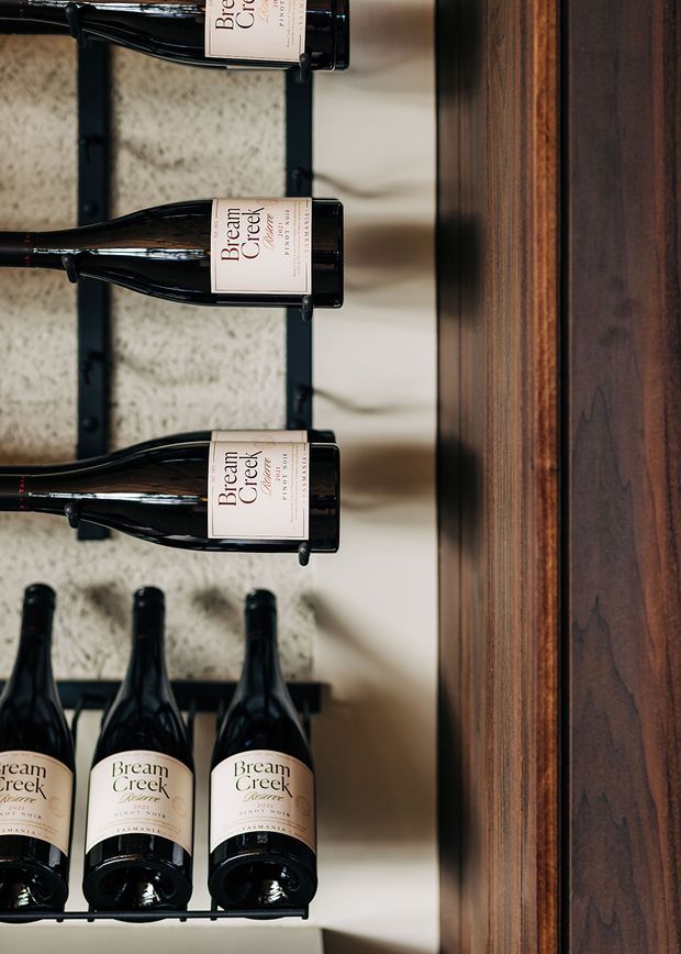 Close-up of a built-in wine storage display with several dark glass wine bottles held in black metal racks. The bottles are arranged against a textured light wall, beside a substantial dark timber vertical panel or cabinet surround. The image is a detail view focused on wine storage and rich woodwork.