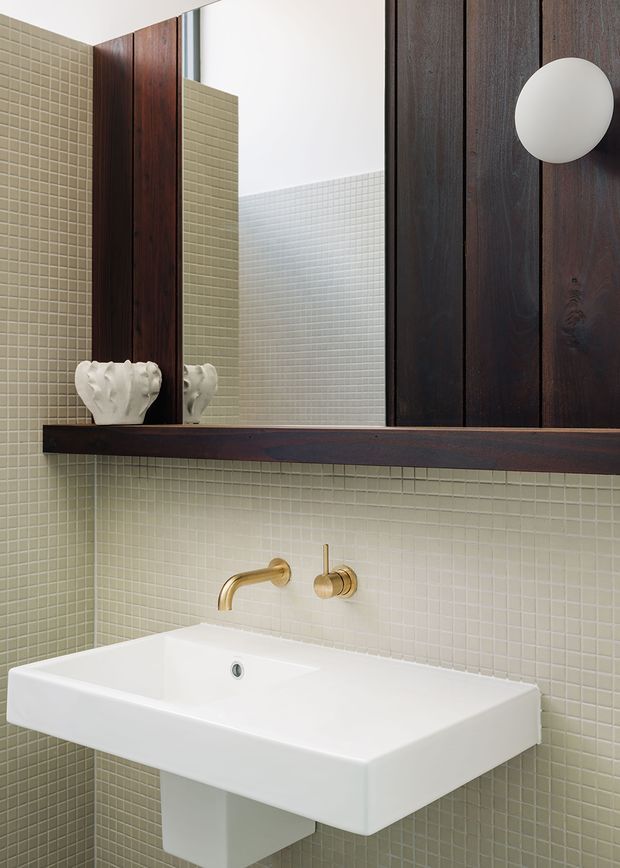 A close-up of a refined bathroom vanity with a wall-mounted white rectangular basin, brushed brass wall-mounted tapware, and pale green small-format mosaic tile covering the walls. A large mirror is framed and supported by dark stained timber wall panelling and a thick timber shelf, with a round white wall light and a decorative white vessel adding detail.