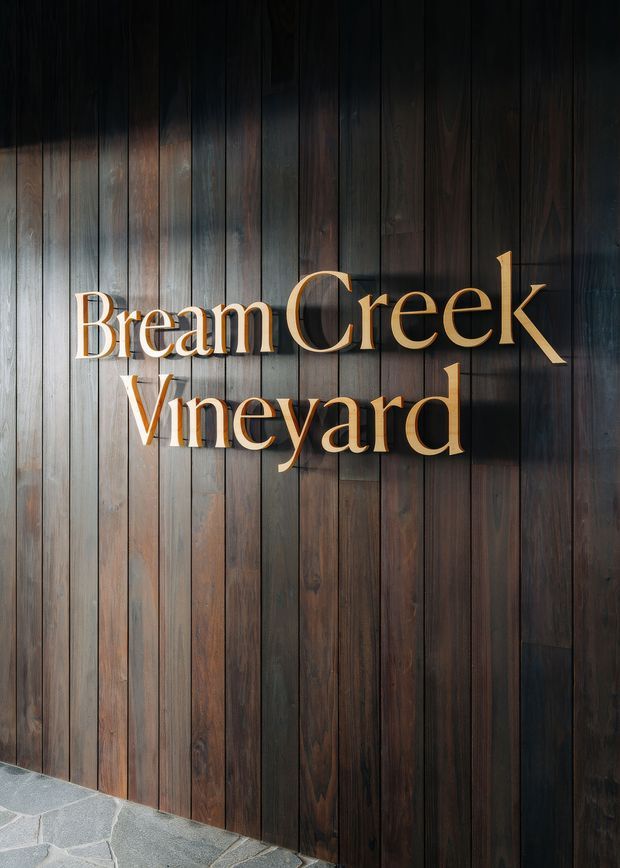 Close-up view of a dark vertical timber-clad wall with prominent raised lettering reading “Bream Creek Vineyard.” A grey irregular stone-paved surface is visible at the base. The image functions as architectural signage or an entrance feature for a vineyard venue.