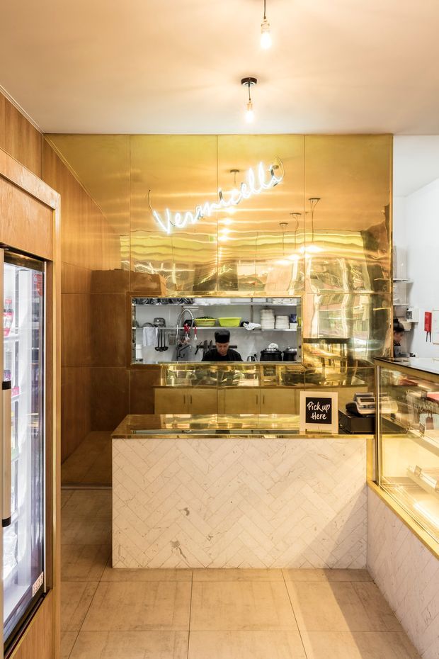 A compact commercial food-service counter viewed from the customer side. The space features extensive reflective brass-gold wall and counter cladding, warm timber wall panelling, pale veined stone-faced cabinetry, tiled flooring, pendant and exposed-bulb lighting, a service opening into a visible commercial kitchen, and a refrigerated display at the left.