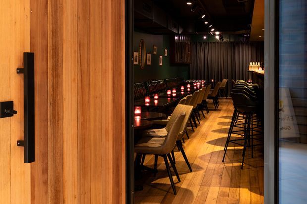A dimly lit contemporary hospitality interior viewed through a large timber pivot door. The space appears to be a restaurant or bar, with a long dark dining counter lined with small glowing lights, upholstered dining chairs, bar stools, dark walls, framed artwork, curtains, and warm timber flooring. Black ceilings with recessed and track lighting create an intimate, dramatic atmosphere.