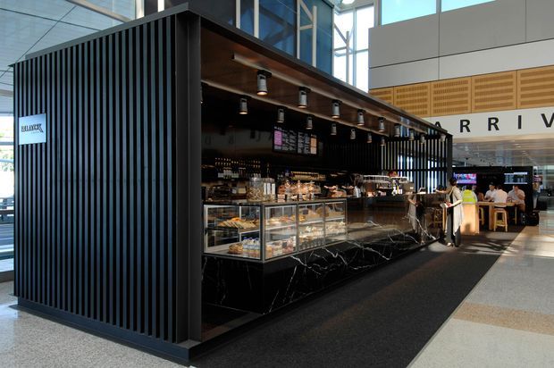 A contemporary café kiosk in a bright, spacious commercial interior. The freestanding café features a dramatic black vertically slatted enclosure, dark marble-look counter fronts, glass pastry display cases, a timber-lined canopy, pendant spot lighting, and an open service counter. Customers are seated in the surrounding atrium or dining area, with large windows and high ceilings creating an airy setting.