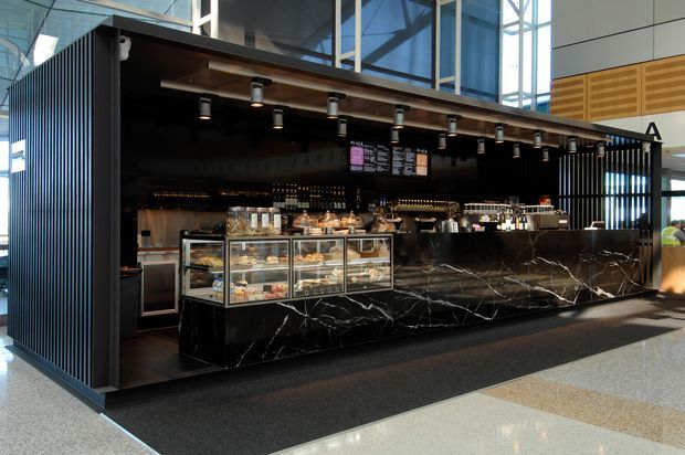 A sleek contemporary café or food-service kiosk in a bright commercial interior. The open counter features dramatic black marble-look cladding with white veining, glass refrigerated display cases, dark timber overhead detailing, black vertical slatted screens, pendant spotlights, and extensive blue-tinted structural glazing behind. The space has a polished, high-contrast hospitality fit-out.